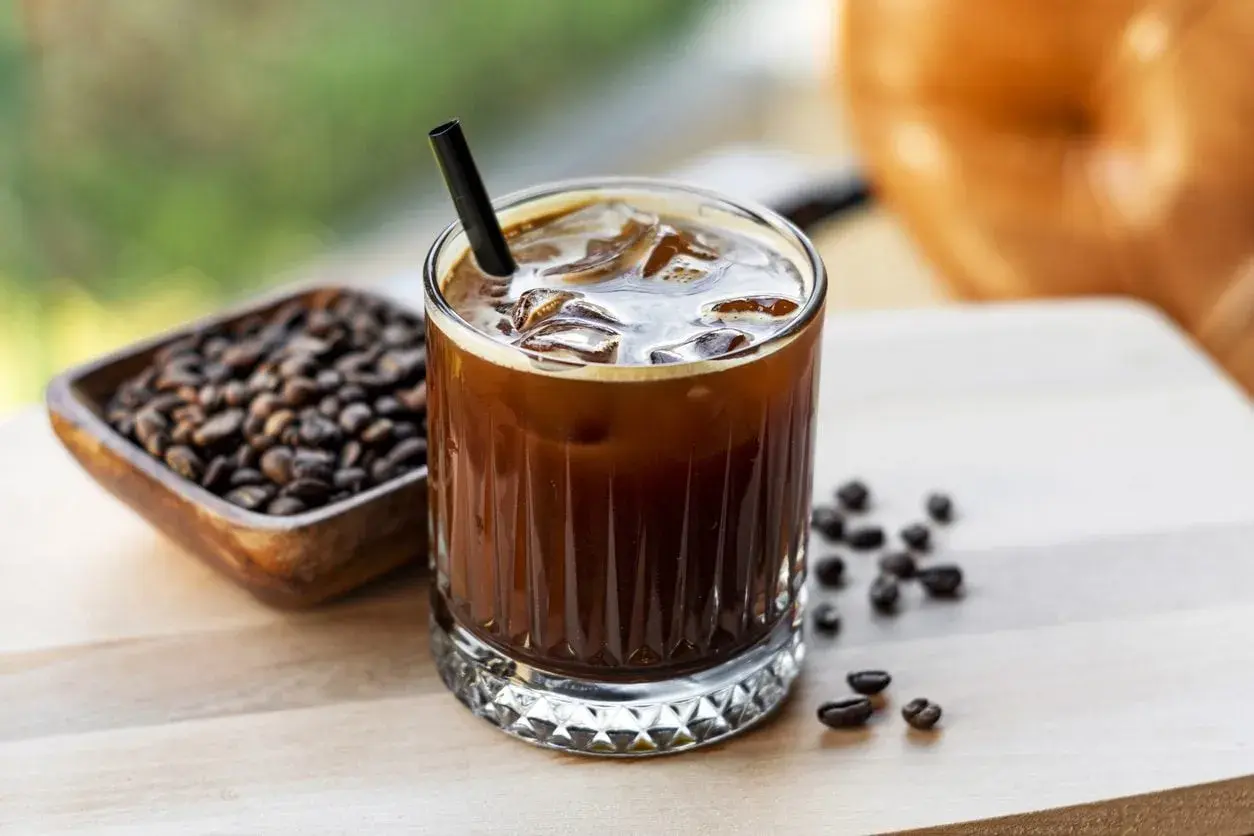 Un bicchiere di **cold brew latte** con ghiaccio e una cannuccia nera, accanto a una ciotola di chicchi di caffè tostati.