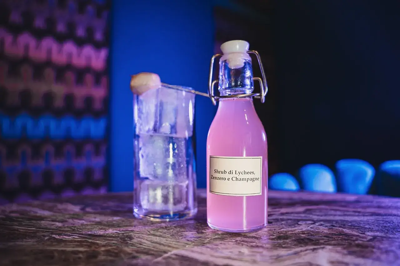 Bottiglia di shrub cocktail rosa con etichetta "Lychees, Zenzero e Champagne" accanto a un bicchiere pieno di ghiaccio.