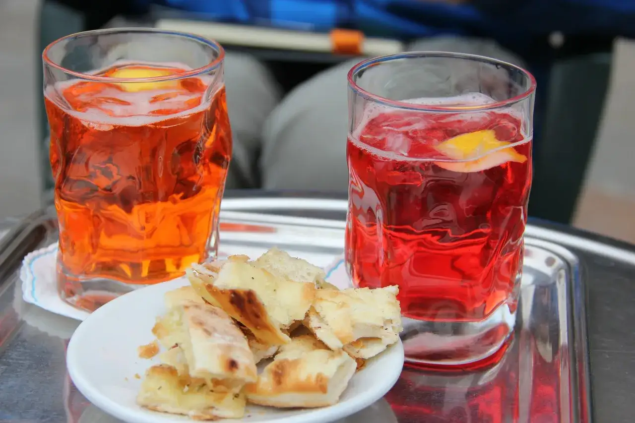 Due bicchieri di aperitivo, uno arancione e uno rosso, con fette di limone e un piattino di focaccia. Un aperitivo originale perfetto per un momento di relax.
