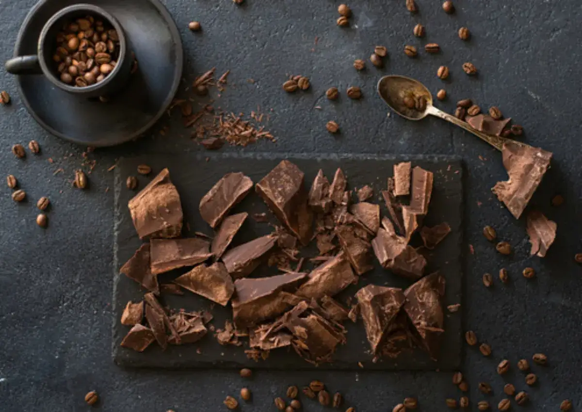 Tavoletta di cioccolato spezzata su ardesia, chicchi di caff&egrave; sparsi e una tazzina piena. Un invito a mangiare chicchi di caff&egrave; e cioccolato.