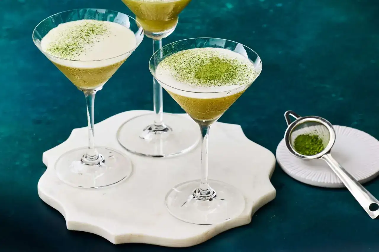 Tre cocktail verdi in coppe da martini, guarniti con polvere di matcha. Un colino con matcha e una base di marmo completano gli ingredienti per un drink giapponese.