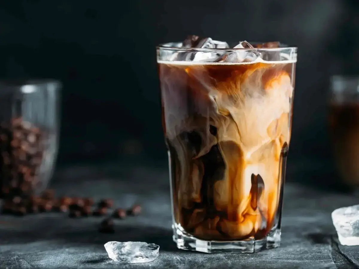 Un bicchiere di latte macchiato freddo con striature di latte e caff&egrave;, cubetti di ghiaccio e chicchi di caff&egrave; sullo sfondo.