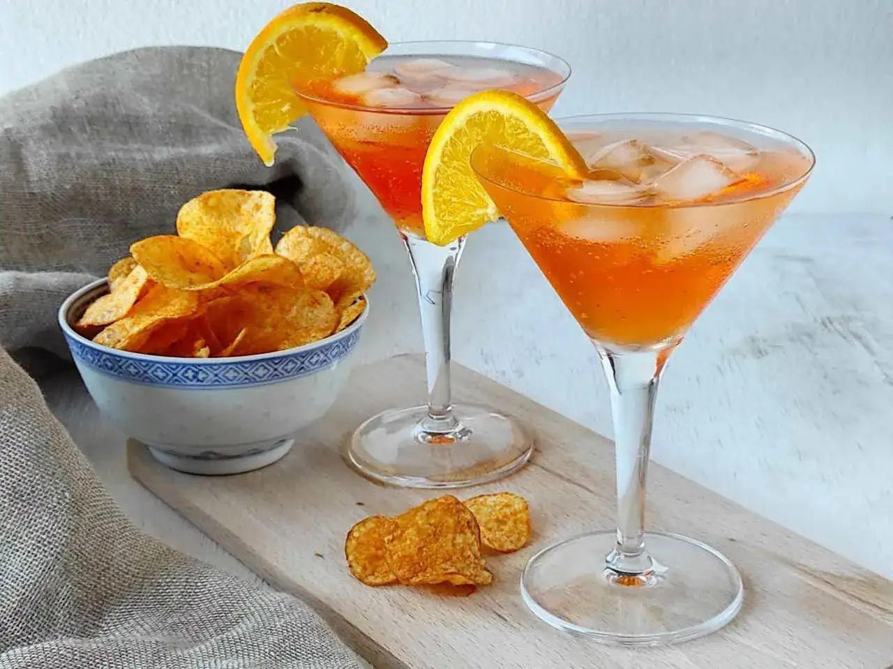 Aperol soda ricetta: due bicchieri di cocktail arancione con ghiaccio e fettine d'arancia, accompagnati da una ciotola di patatine croccanti.