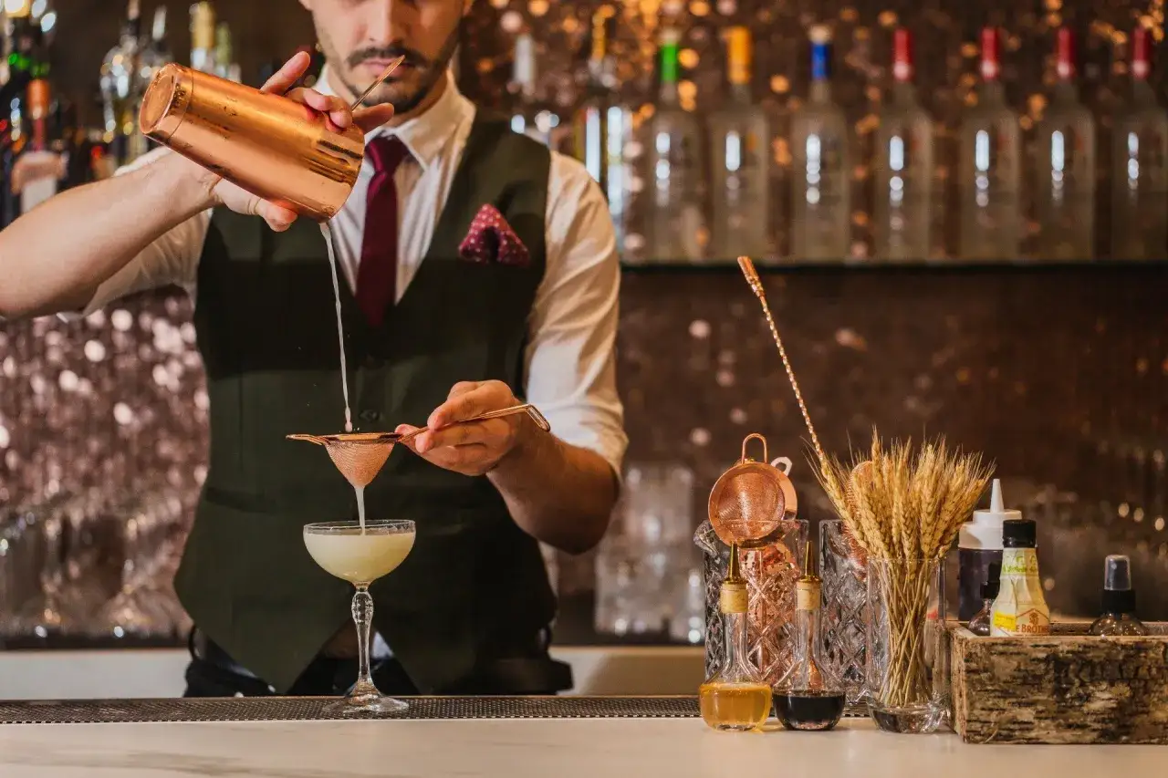 Barman esperto prepara cocktail difficili, filtrando un drink in una coppa elegante. Dietro, bottiglie illuminate e spighe di grano.