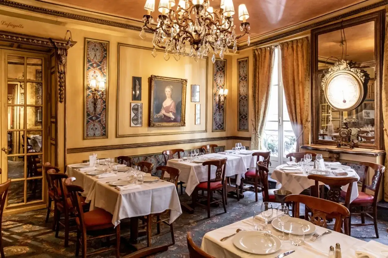 Interno elegante di uno dei locali storici di Parigi, con tavoli apparecchiati, un grande lampadario e un ritratto d'epoca.