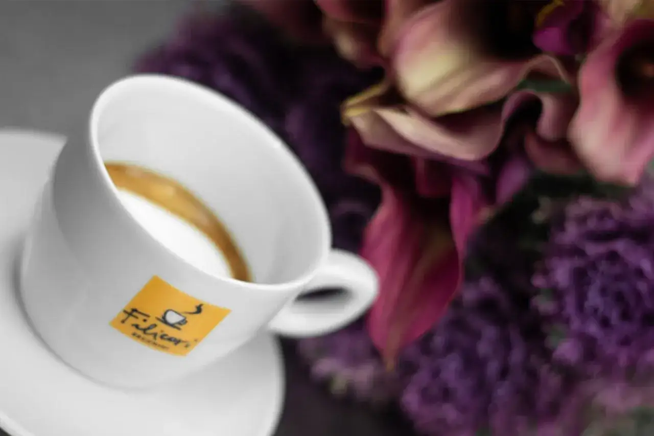 Tazza di caffè con schiuma e logo Filicori. Un macchiatone o un macchiato? Accanto, fiori viola sfocati.