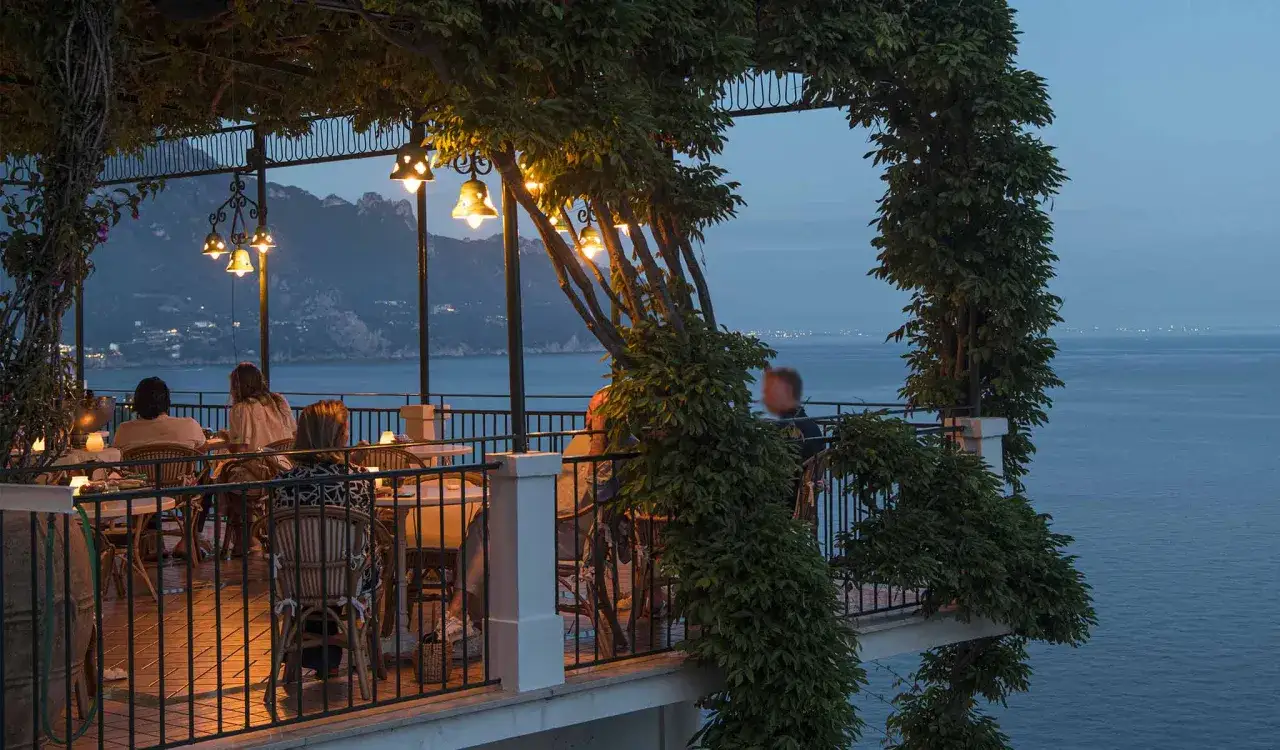 Persone gustano un caffè al mare, seduti in un pergolato illuminato da lanterne, con vista su montagne e mare al crepuscolo.
