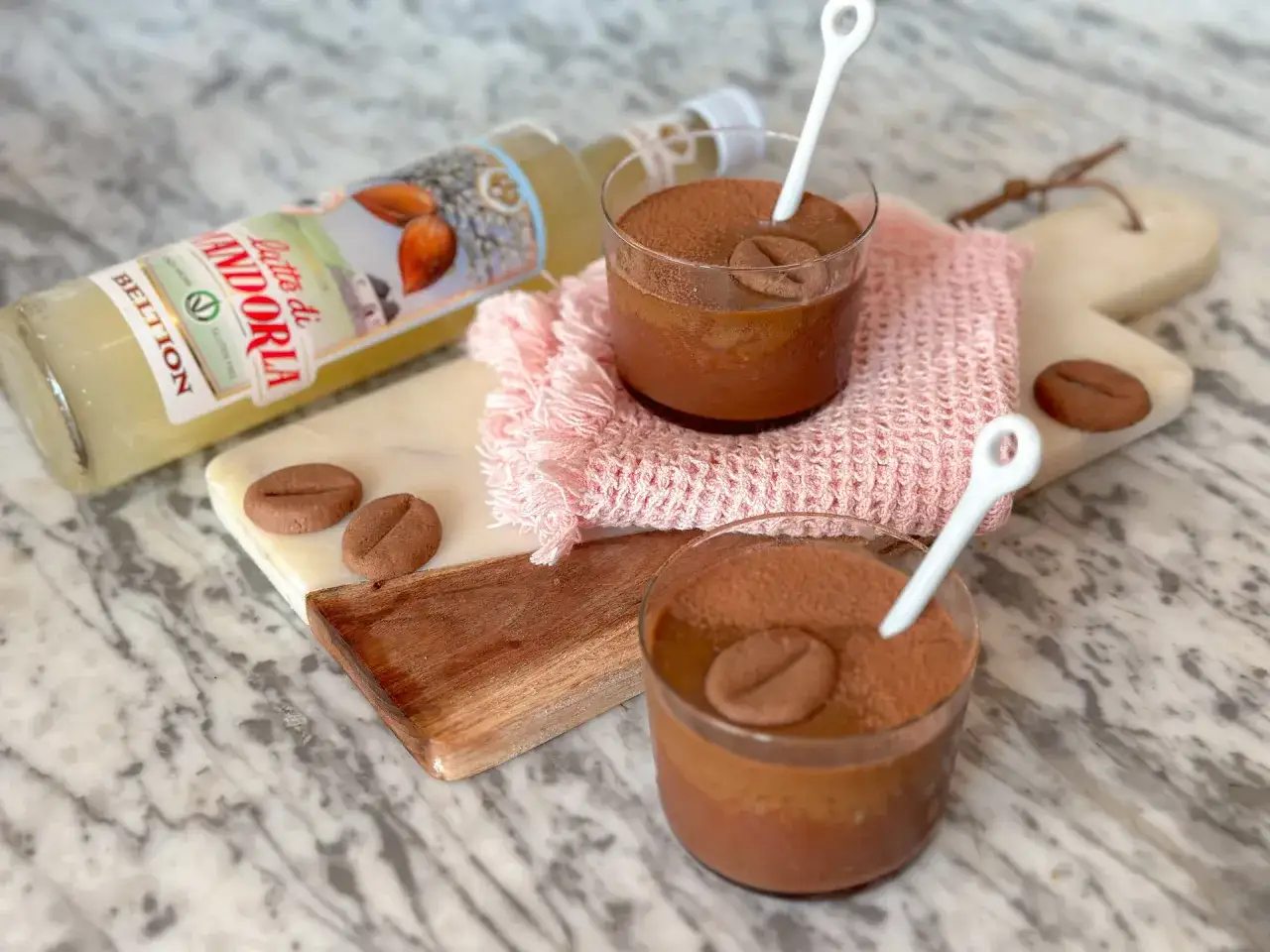 Due bicchierini di mousse al cioccolato con cacao e biscotti a forma di chicco di caffè, accanto a una bottiglia di latte di mandorla.