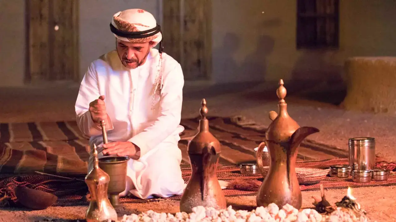 Uomo in abito tradizionale prepara il caff&egrave; arabo pestando spezie in un mortaio, circondato da caffettiere e braci ardenti.