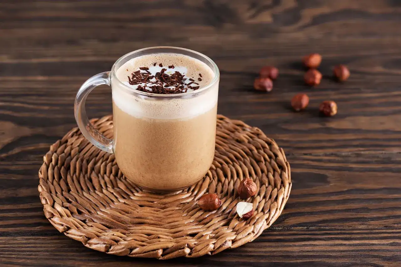 Un bicchiere di mocha coffee con schiuma e scaglie di cioccolato, su un sottobicchiere di paglia, con nocciole sparse.
