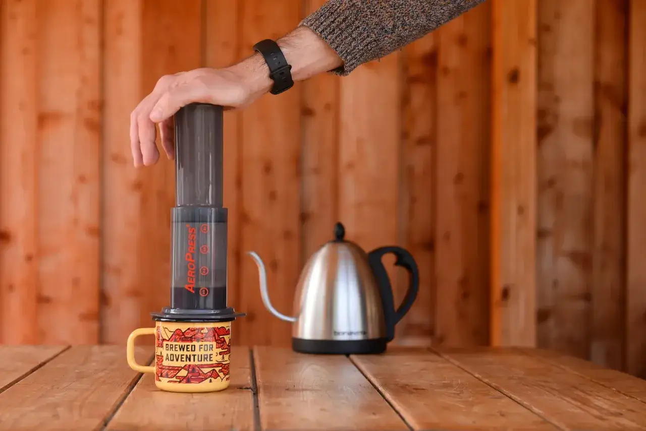 Mano che preme l'Aeropress caff&egrave; su una tazza gialla, con bollitore in acciaio sullo sfondo.