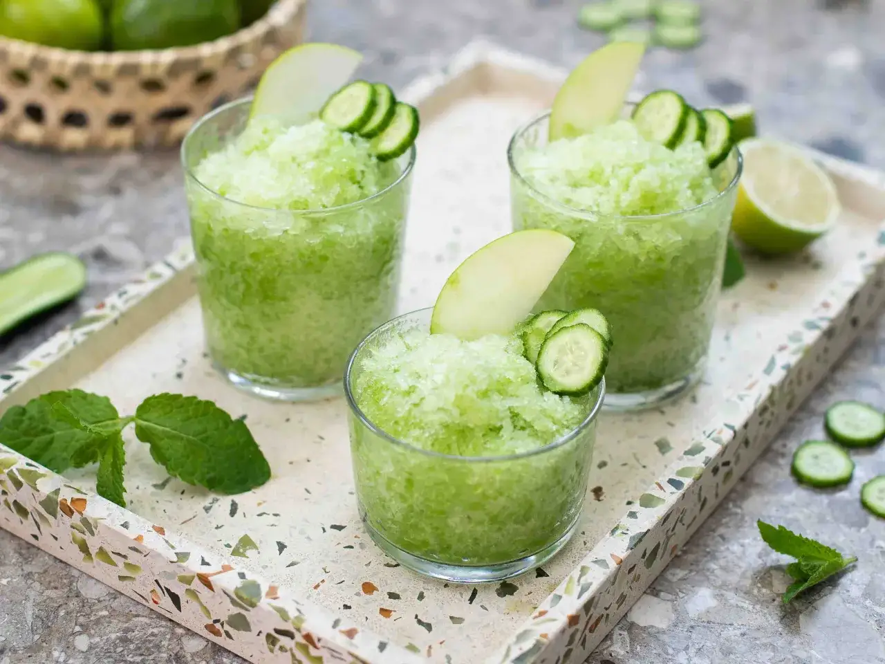 Tre bicchieri di granita verde brillante, guarniti con fettine di mela e cetriolo, perfetti per le bevande estive.
