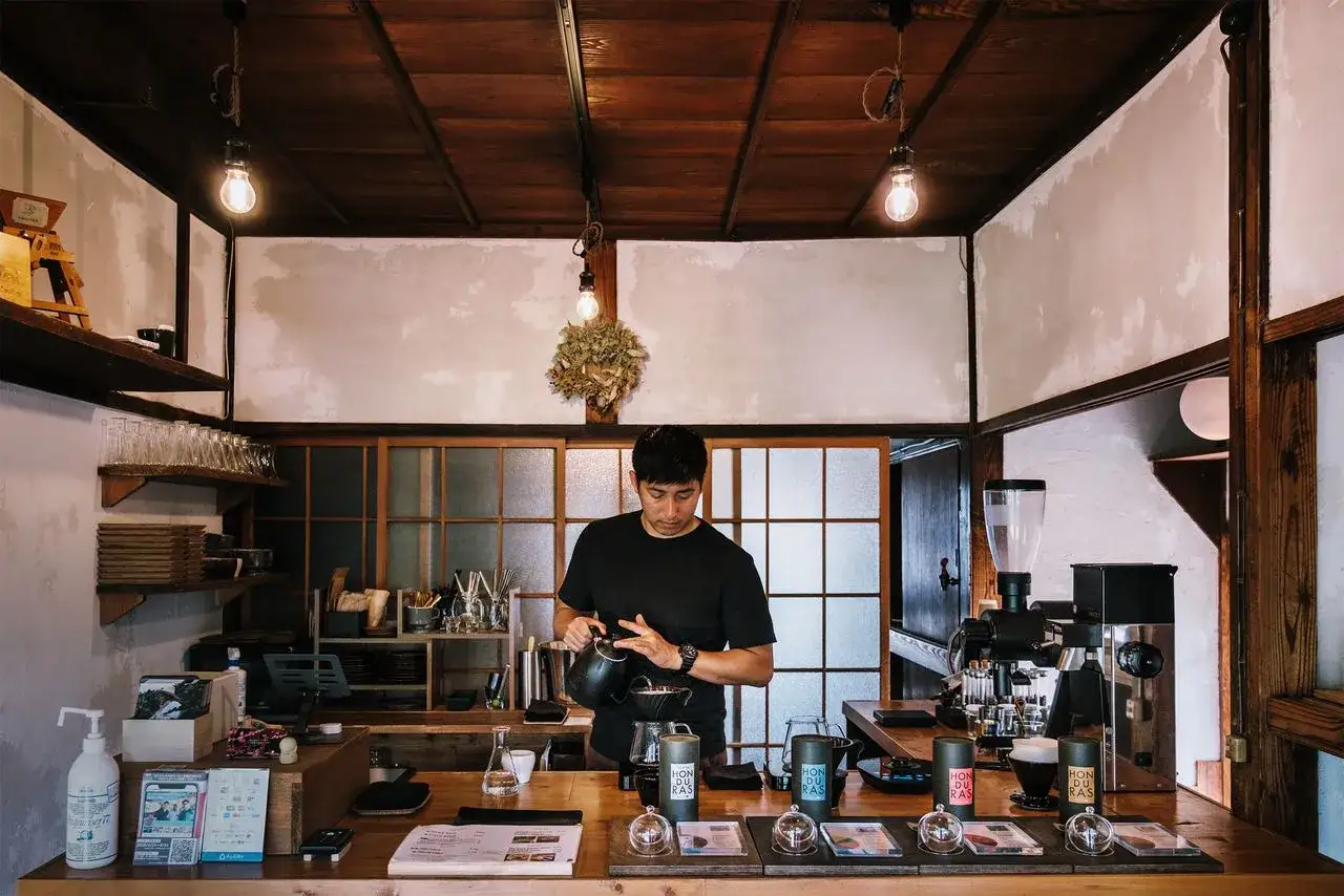 Barista prepara un caffè giapponese in un locale accogliente con soffitto in legno e luci soffuse.