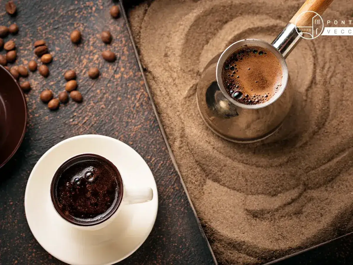 Un caff&egrave; arabo fumante in una tazzina bianca, accanto a un cezve su sabbia calda. Chicchi di caff&egrave; sparsi.