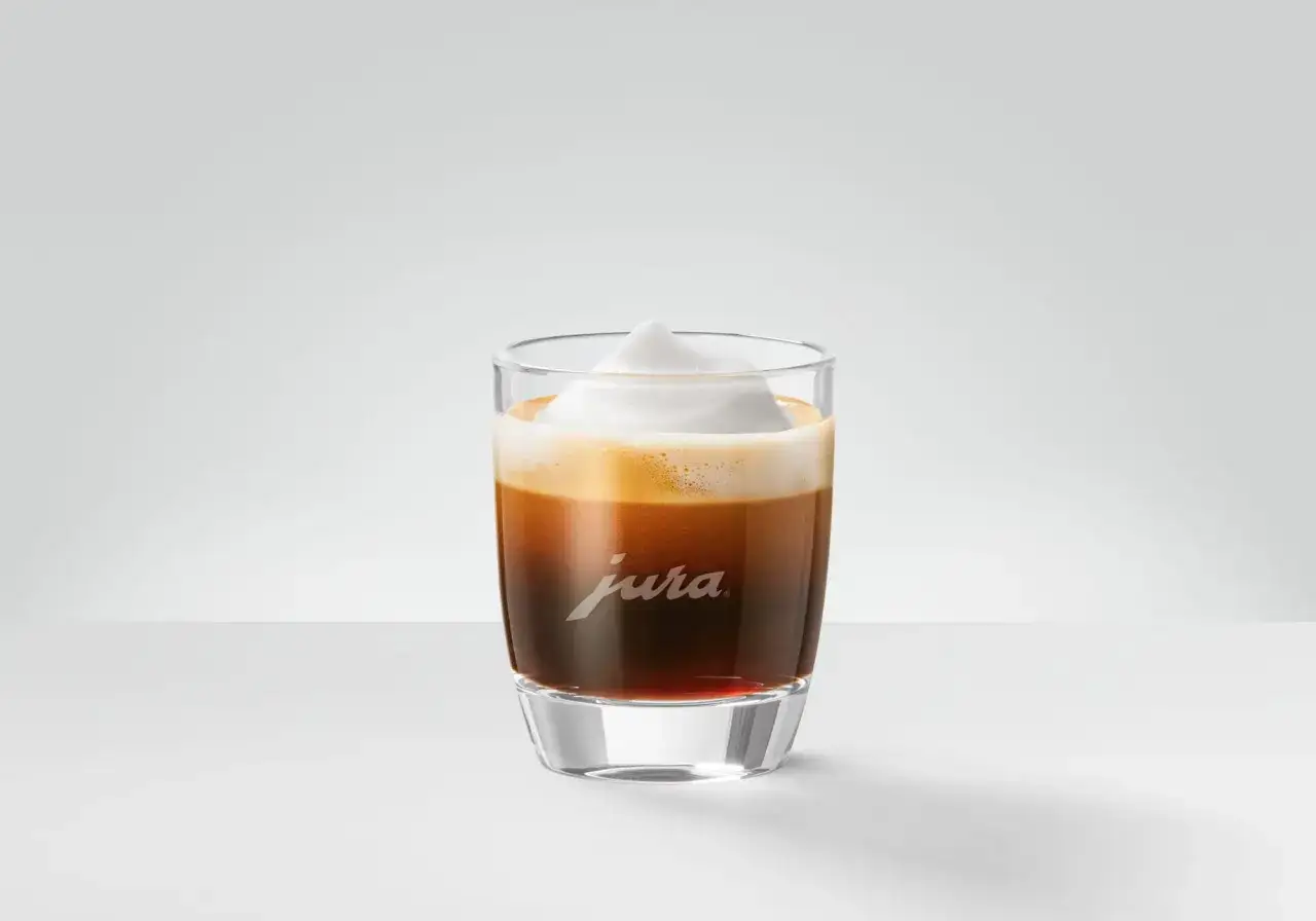 Un bicchiere di caff&egrave; macchiato con schiuma di latte soffice, con il logo Jura inciso sul vetro.