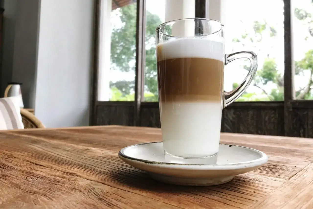 Un caffè macchiato caldo a strati, servito in un bicchiere trasparente su un piattino bianco, su un tavolo di legno rustico.