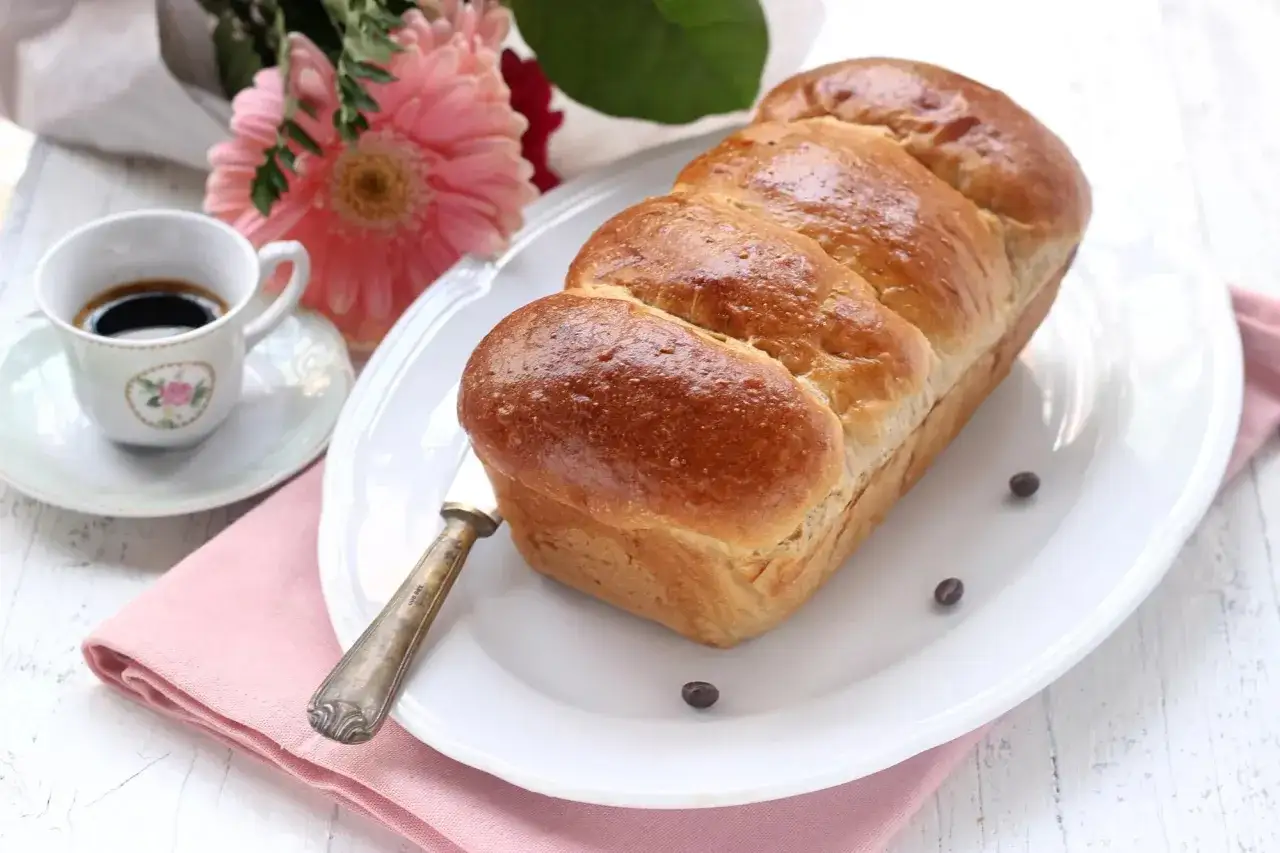 Pane brioche dorato su piatto bianco, accanto a una tazzina di caff&egrave; e fiori rosa. Perfetto per conservare il caff&egrave; avanzato e gustarlo con una fetta.