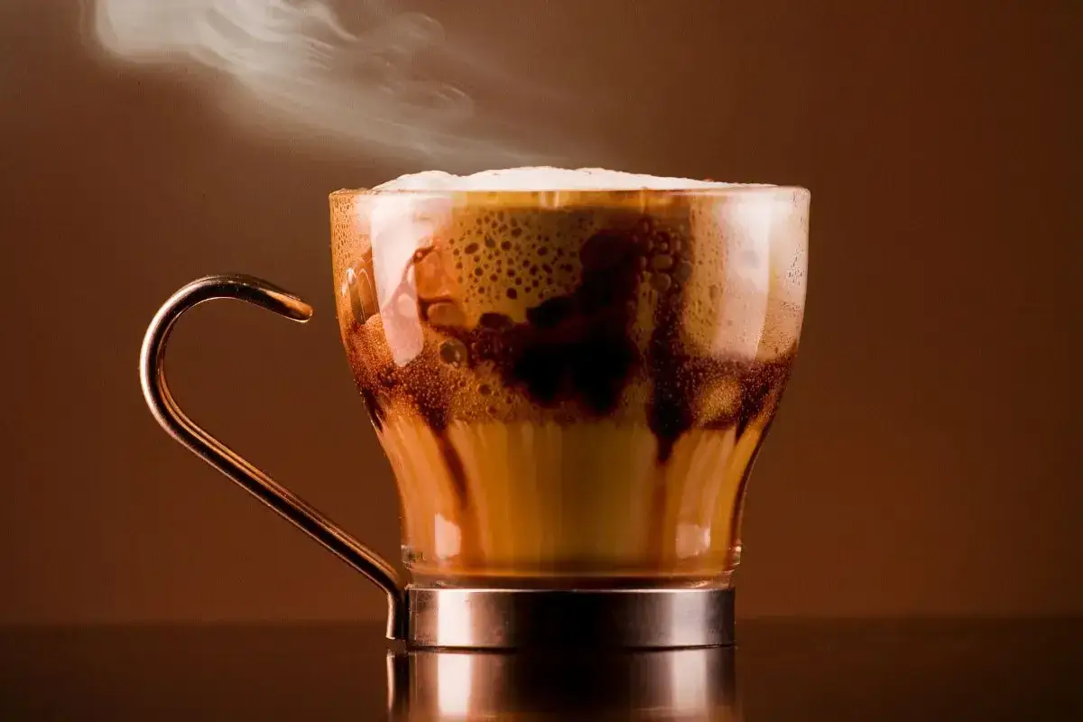 Un fumante caffè marocchino in una tazza di vetro con manico dorato, con schiuma e gocce di cioccolato.