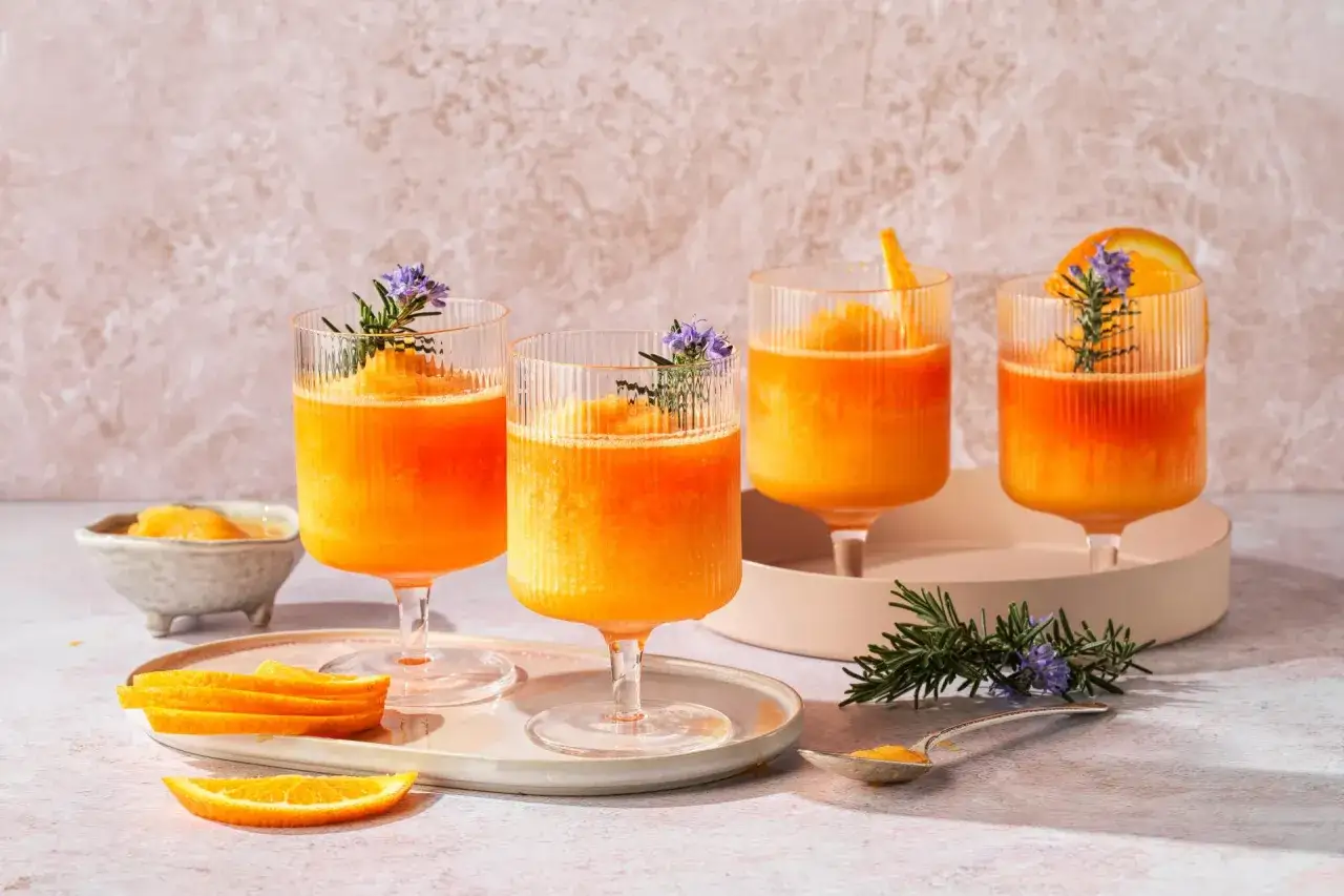 Quattro bicchieri di Aperol Spritz ghiacciato, guarniti con rosmarino e fiori viola, perfetti per una ricetta estiva.