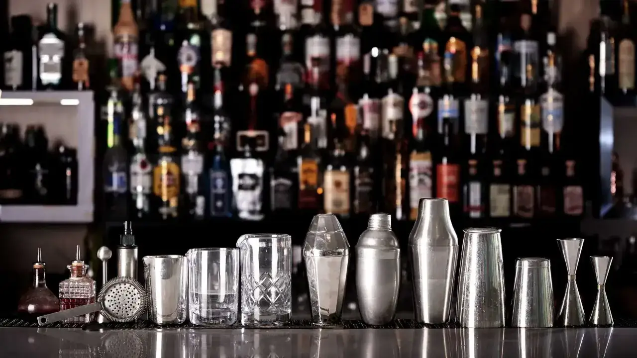 Attrezzatura da bar per preparare una ricetta cocktail: shaker, bicchieri, jigger e strainer, con bottiglie di liquori sullo sfondo.