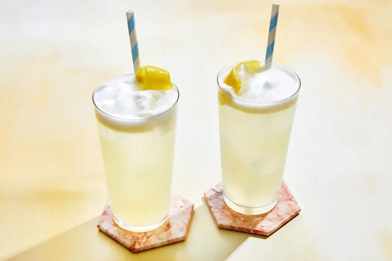 Due cocktail fizz rinfrescanti con schiuma e scorza di limone, serviti su sottobicchieri di marmo.