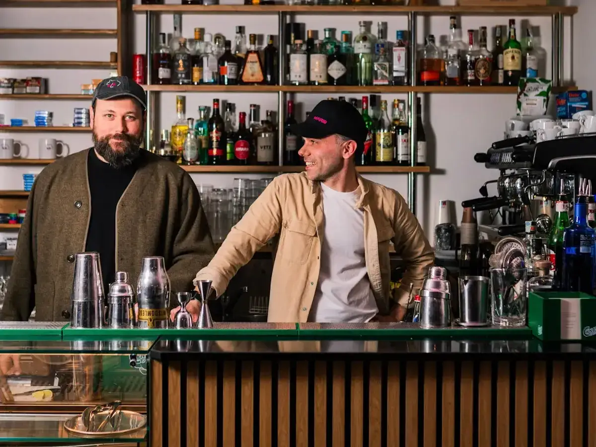 Due baristi sorridono dietro un bancone, pronti per il rito del caff&egrave; e cocktail. Scaffali pieni di bottiglie alle loro spalle.
