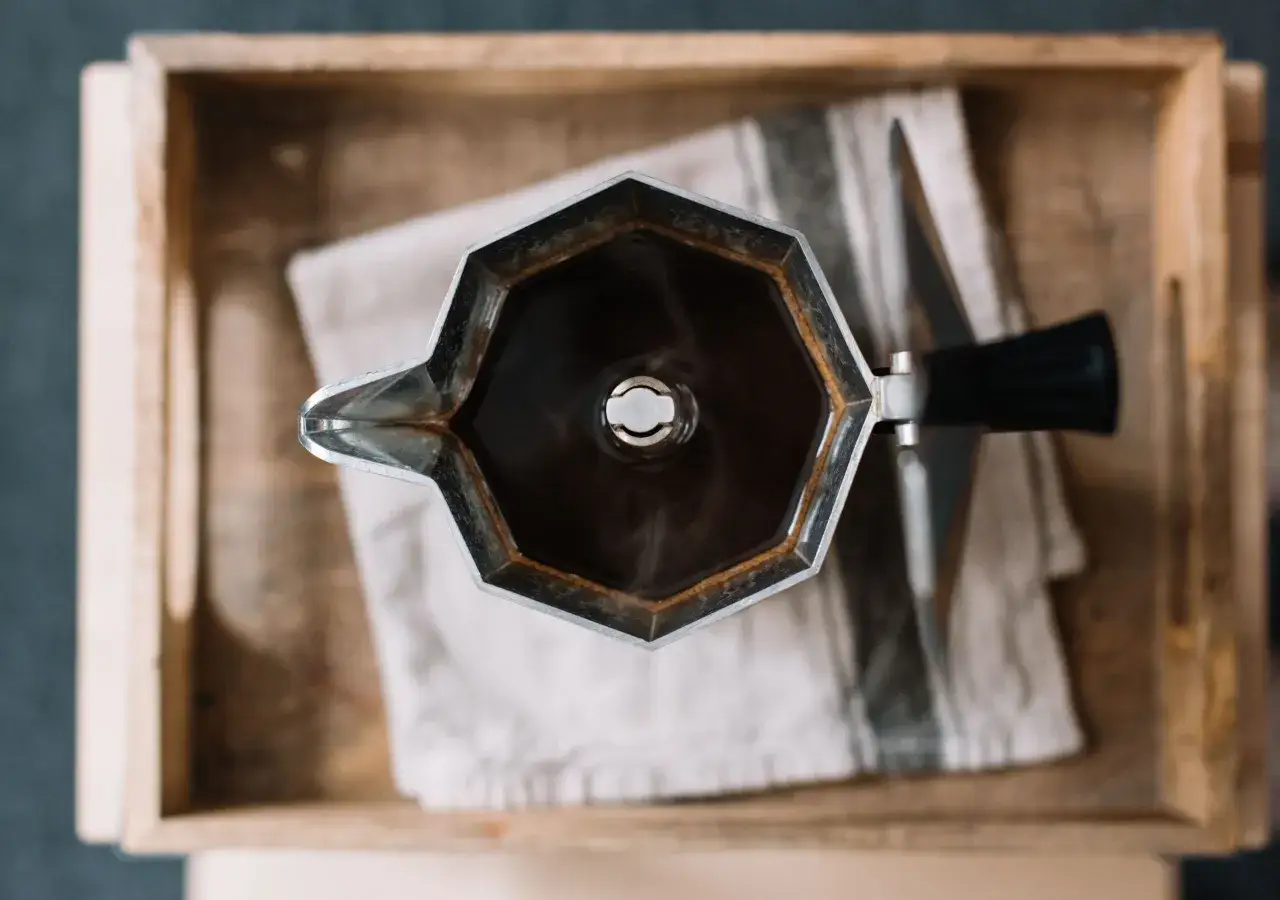 Moka nuova, appena usata, con caffè fumante, appoggiata su un panno a righe dentro una cassetta di legno.