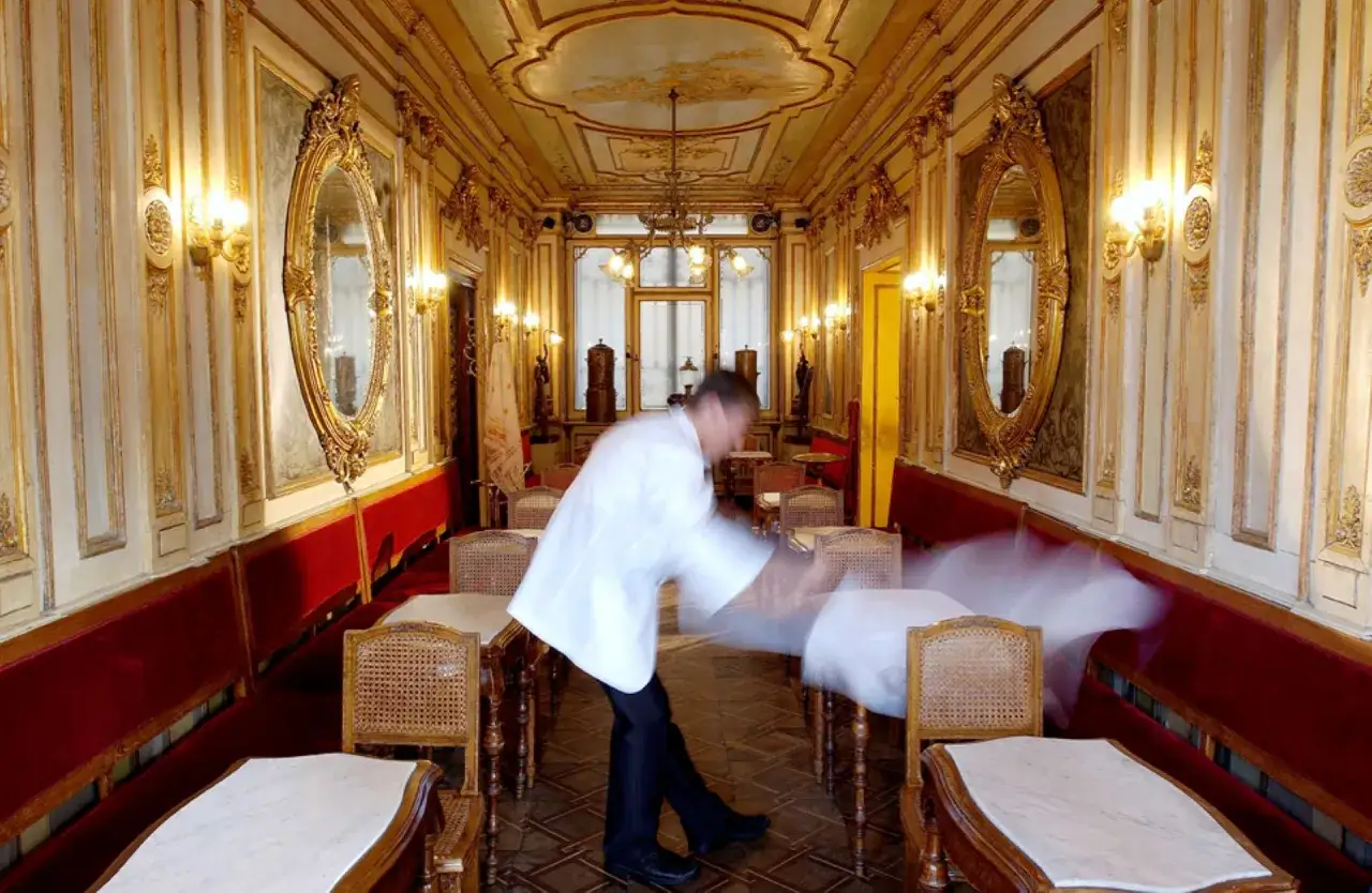 Cameriere in movimento nel lussuoso Cafe Florian, con specchi dorati e sedute rosse.