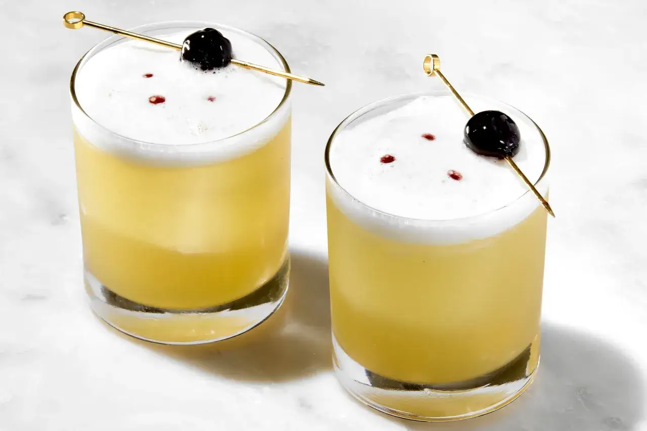 Due cocktail aspri con schiuma bianca, guarniti con ciliegie e stecchini dorati, su sfondo marmorizzato.