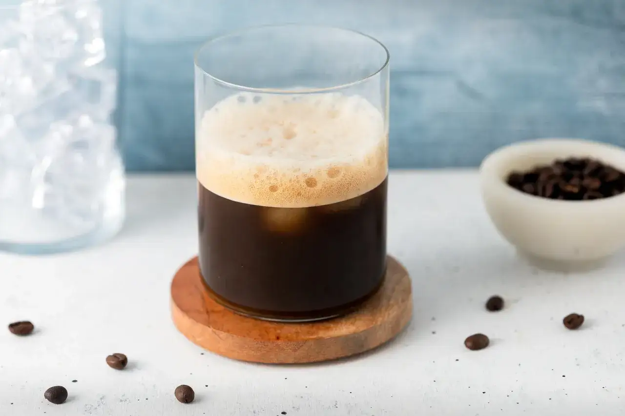 Un bicchiere di caff&egrave; shakerato cremoso con schiuma dorata, cubetti di ghiaccio e chicchi di caff&egrave; tostati.