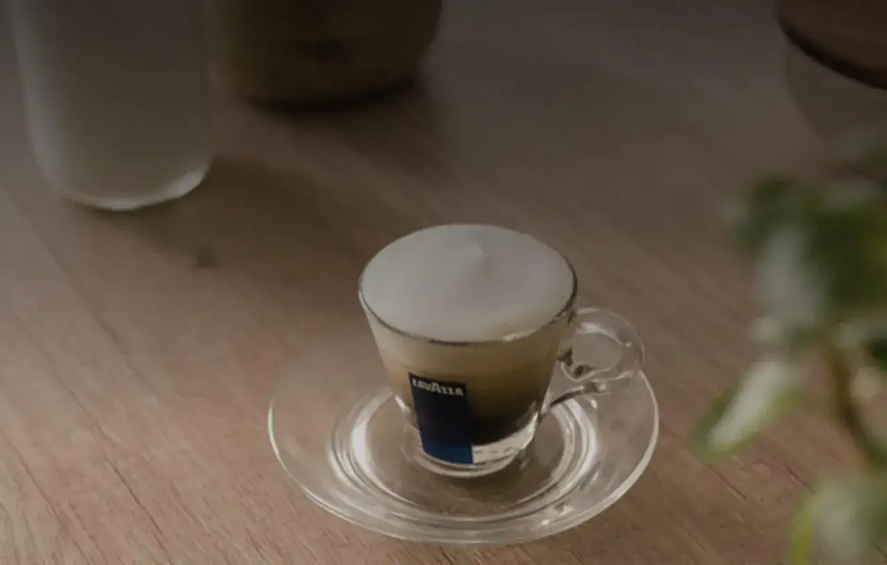 Un macchiatone, o forse un macchiato? Una tazza di caffè con schiuma di latte su un piattino trasparente.