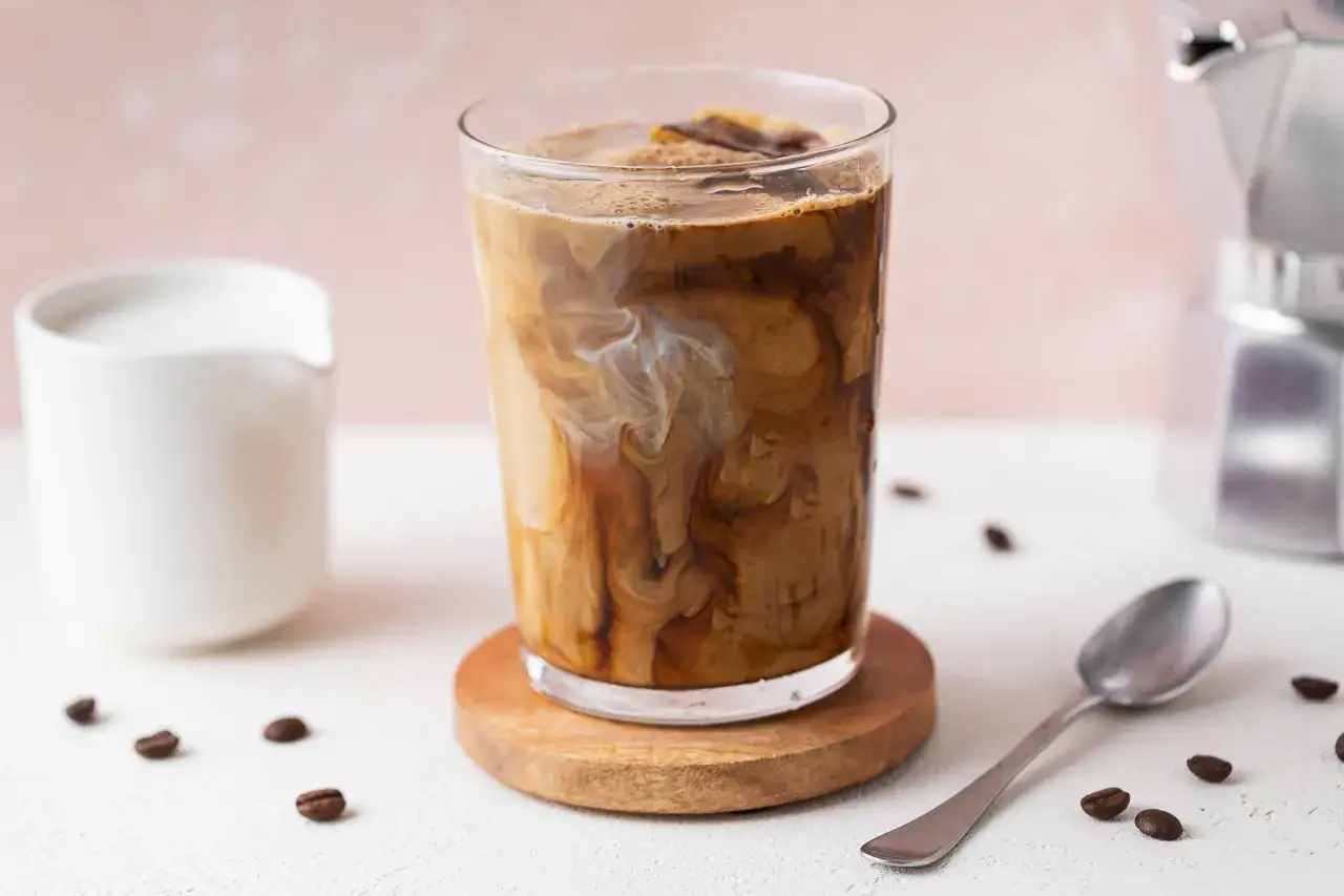 Un bicchiere di iced coffee con latte che crea vortici artistici, su un sottobicchiere di legno. Vicino, una zuccheriera bianca e chicchi di caff&egrave;.