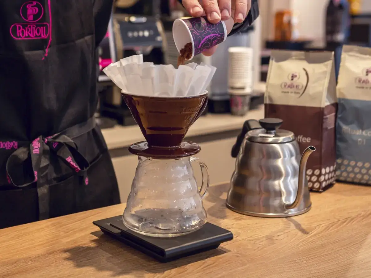 Preparazione di un caffè drip con caffè macinato versato in un filtro. Bollitore e sacchetti di caffè sullo sfondo.