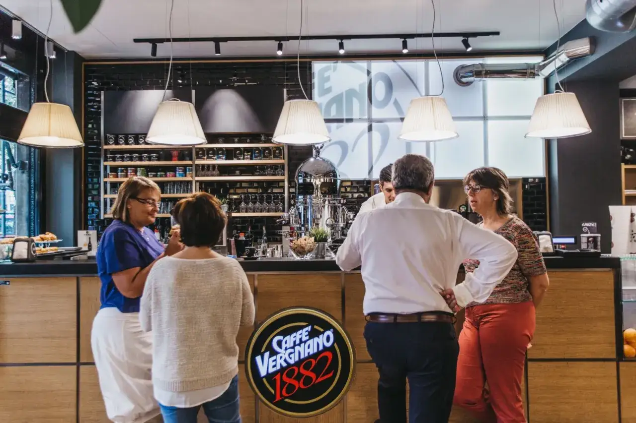 Amici al bar per un caffè, chiacchierando davanti al bancone con il logo Caffè Vergnano 1882.