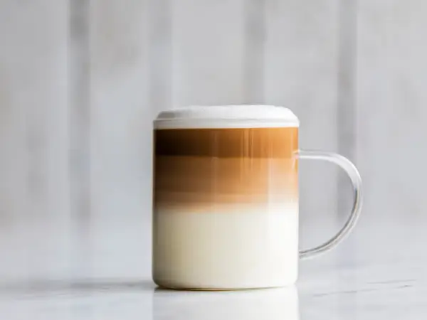 Un bicchiere di latte macchiato freddo con strati distinti di latte, caffè e schiuma, su uno sfondo sfocato.