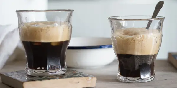 Due bicchieri di caffè shakerato cremoso, con la schiuma dorata che sovrasta il caffè scuro. Uno ha un cucchiaino.